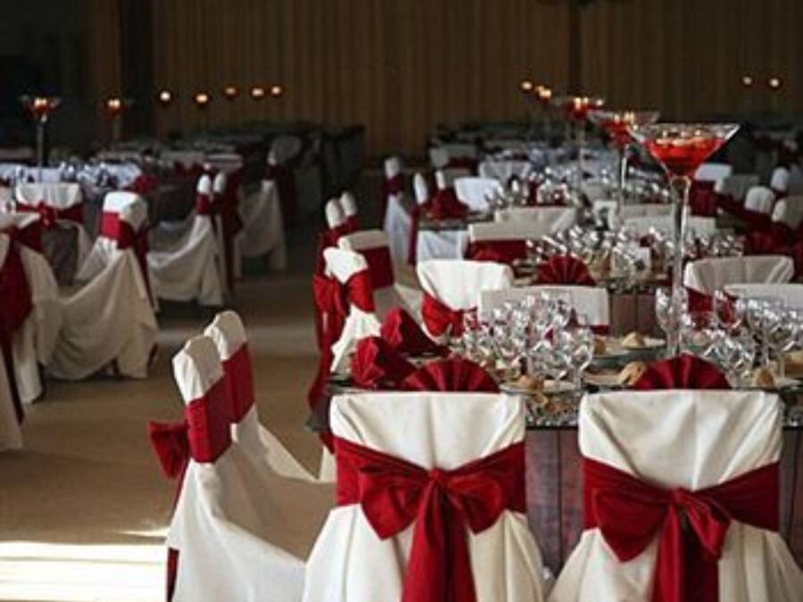 Un salón de banquetes con mesas dispuestas para un evento formal, cada una con un camino de mesa rojo y blanco y una copa de vino tinto.
