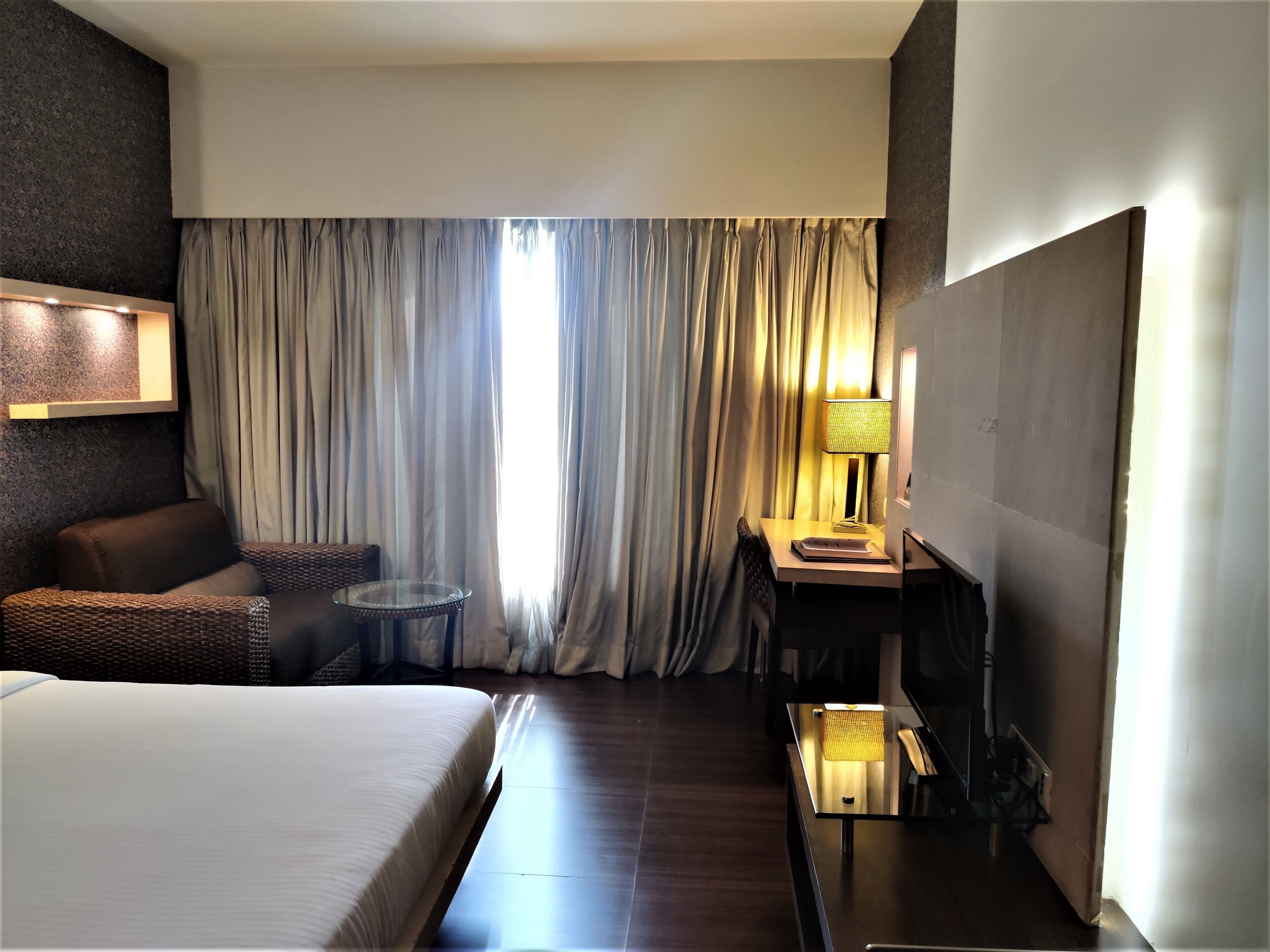 A hotel room with a bed, a desk, a chair, a small table, and a lamp.