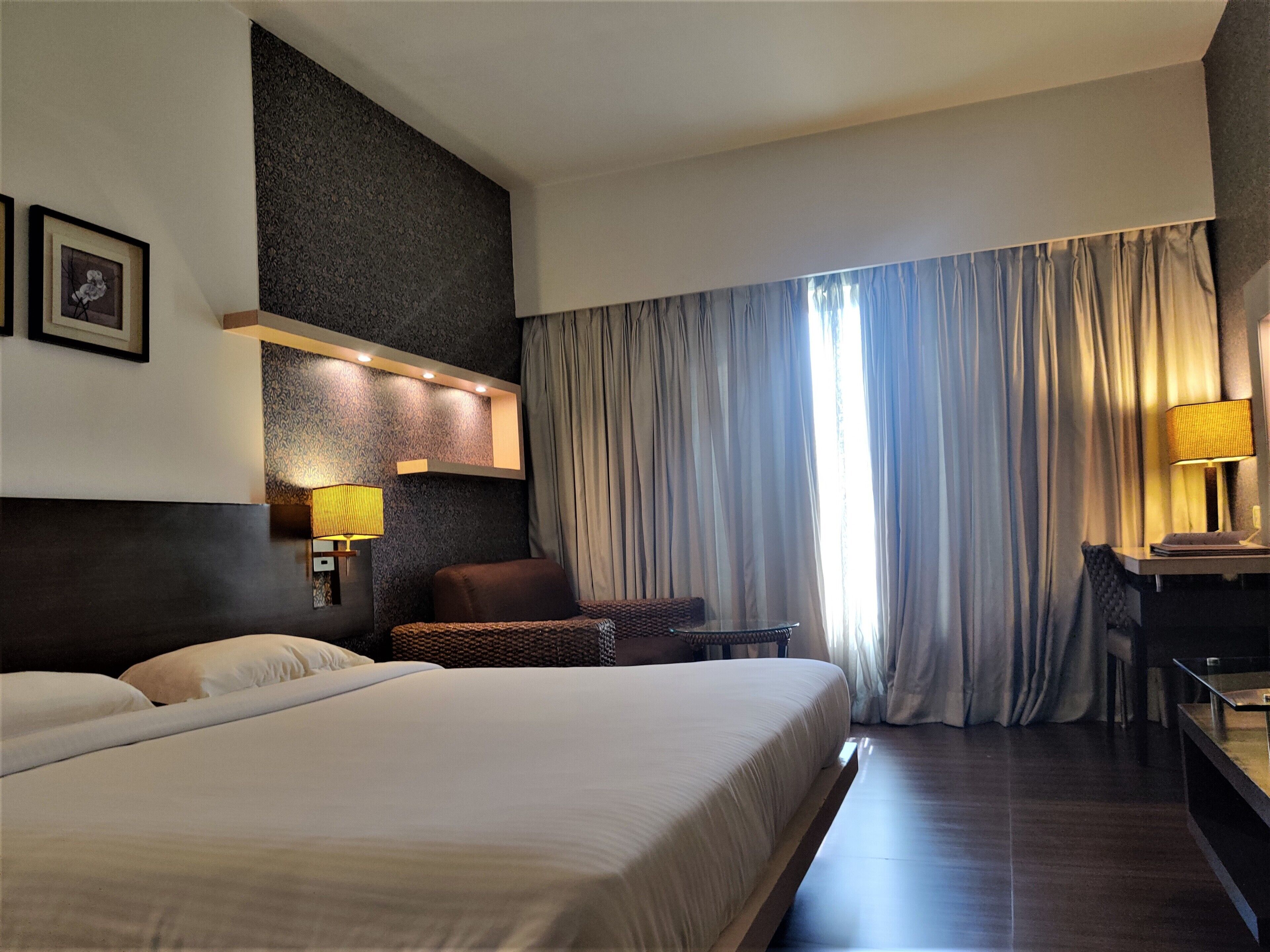 A hotel room with a large bed, a desk with a lamp, a chair, and a sofa. There is a window with curtains and a wall-mounted light fixture.