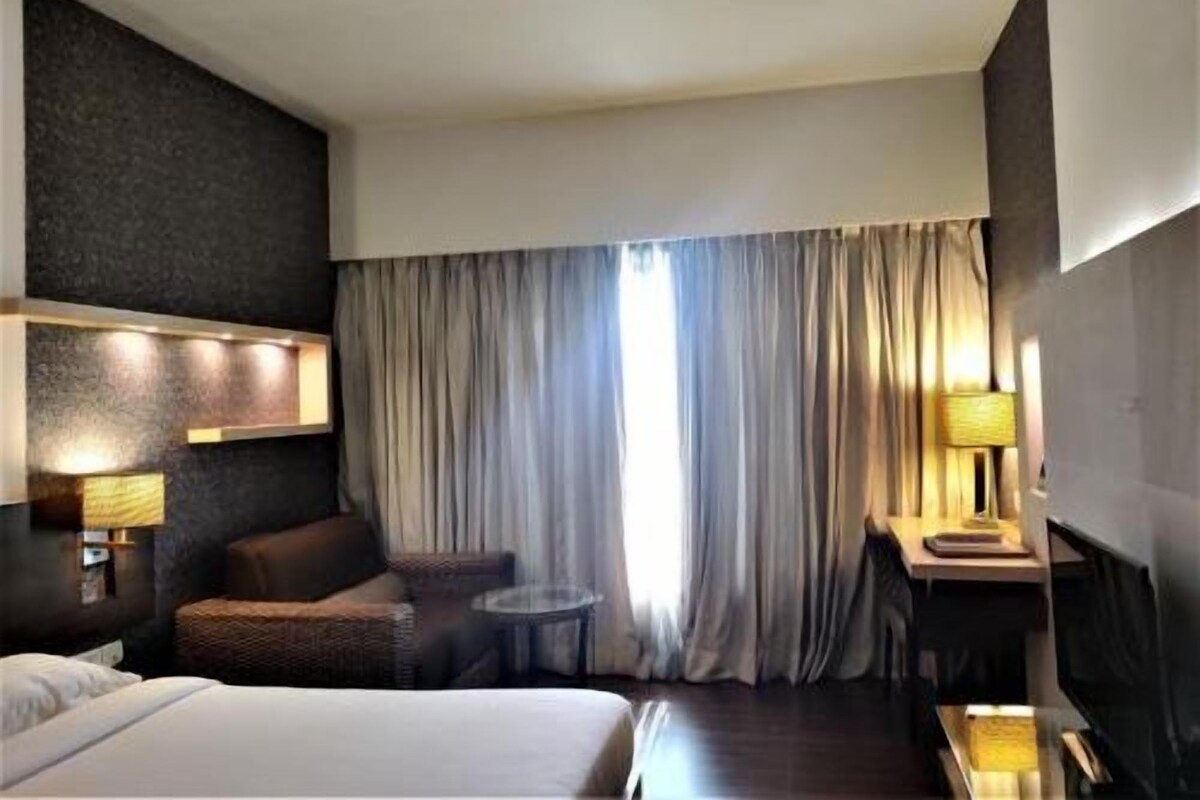 A hotel room with a bed, a sofa, a small table, a lamp, and a large window with curtains.