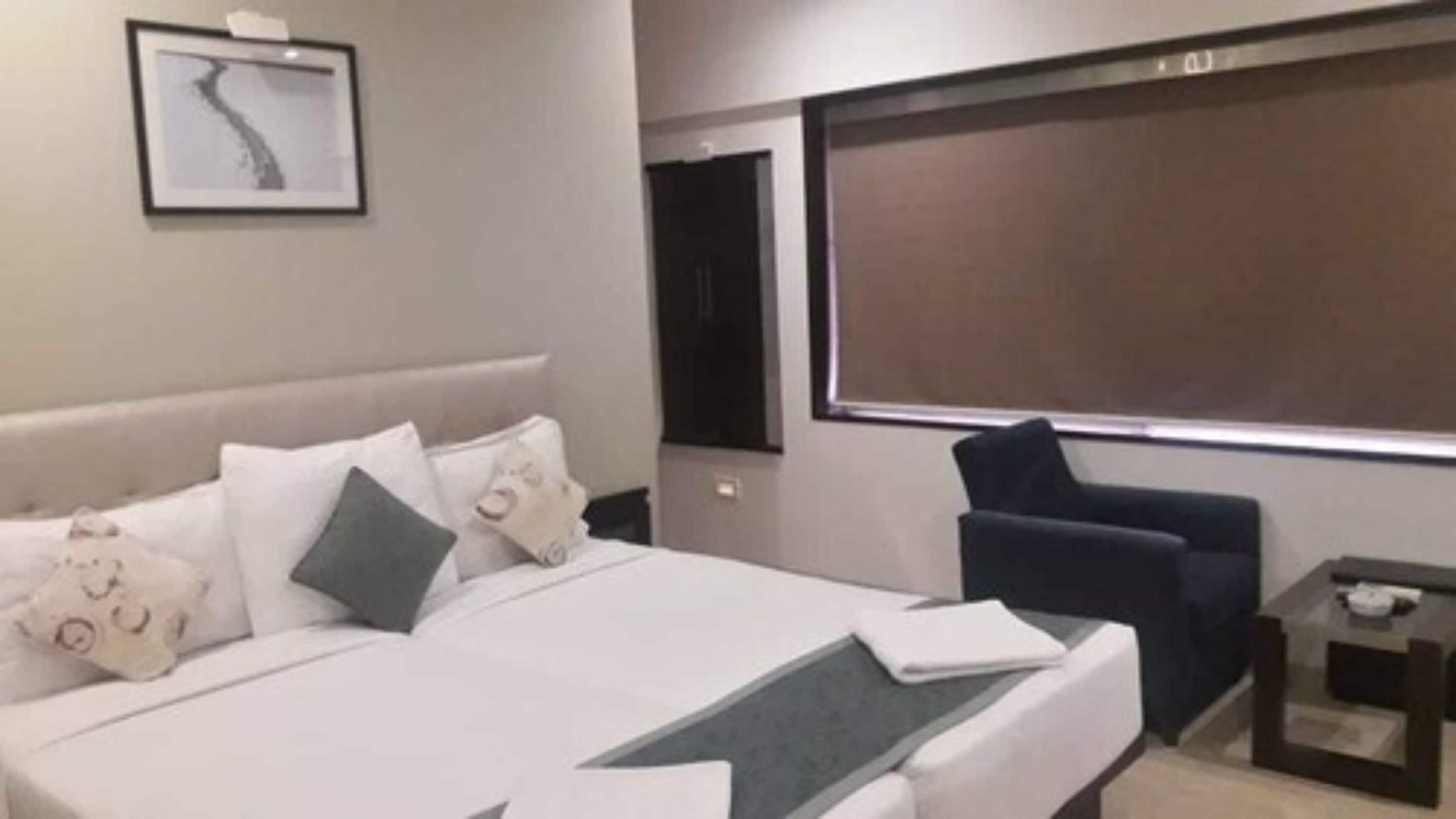 Een hotelkamer met een groot bed, een donkere fauteuil, een klein tafeltje en een groot raam.