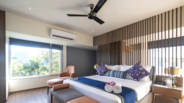 A modern hotel room with a large bed, a bench, a ceiling fan, an air conditioning unit, and a view of the city. - Hotel Le Grande - Mumbai International Airport (Mumbai)