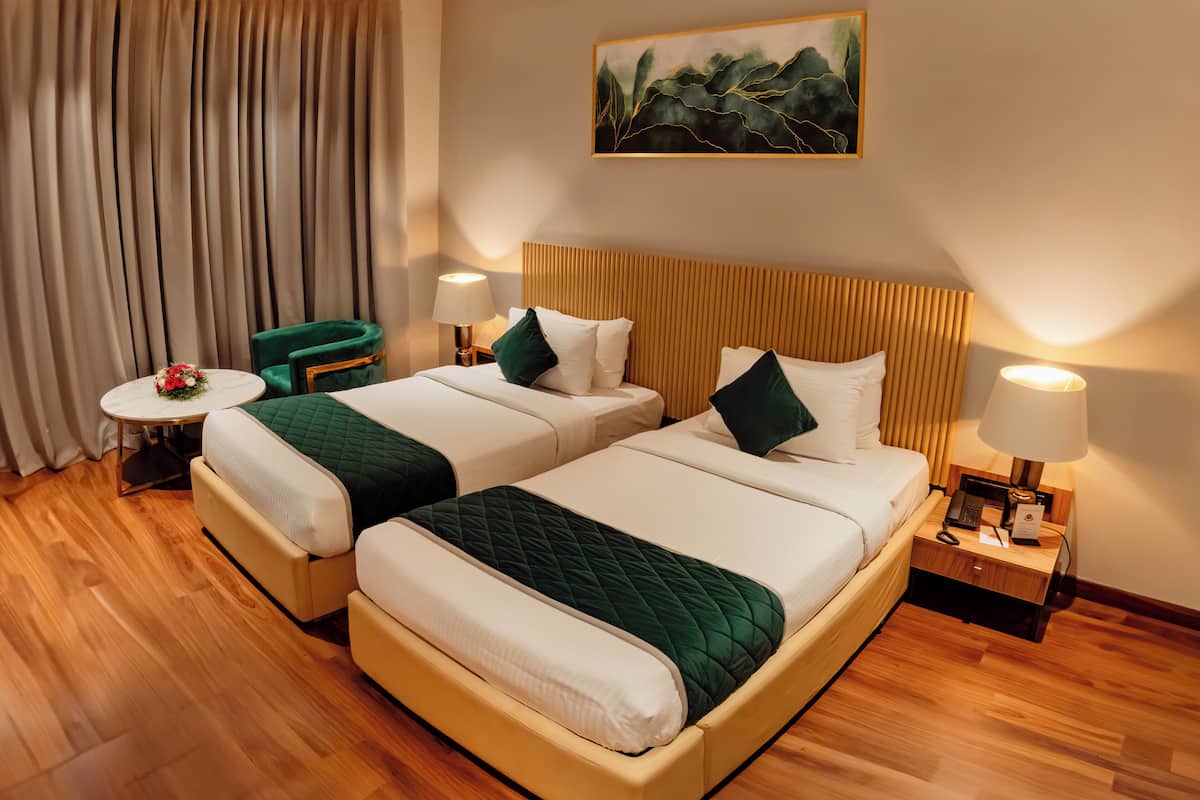 A hotel room with two beds, a small table with a vase of flowers, a green chair, and a painting on the wall.