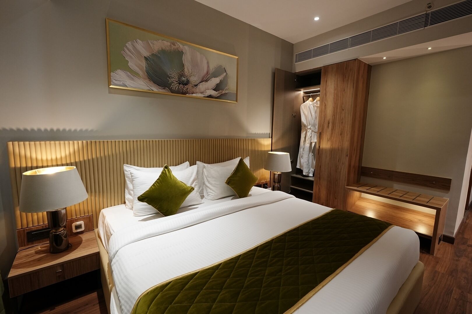 A hotel room with a large bed, two bedside lamps, a wooden headboard, a green quilted bedspread, a wall-mounted artwork, and a walk-in closet with a robe.