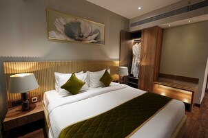 Quarto de hotel com uma cama grande, duas luminárias de cabeceira, um painel de cabeceira de madeira, um edredom acolchoado verde e uma obra de arte pendurada na parede. Possui também um closet espaçoso com roupão.