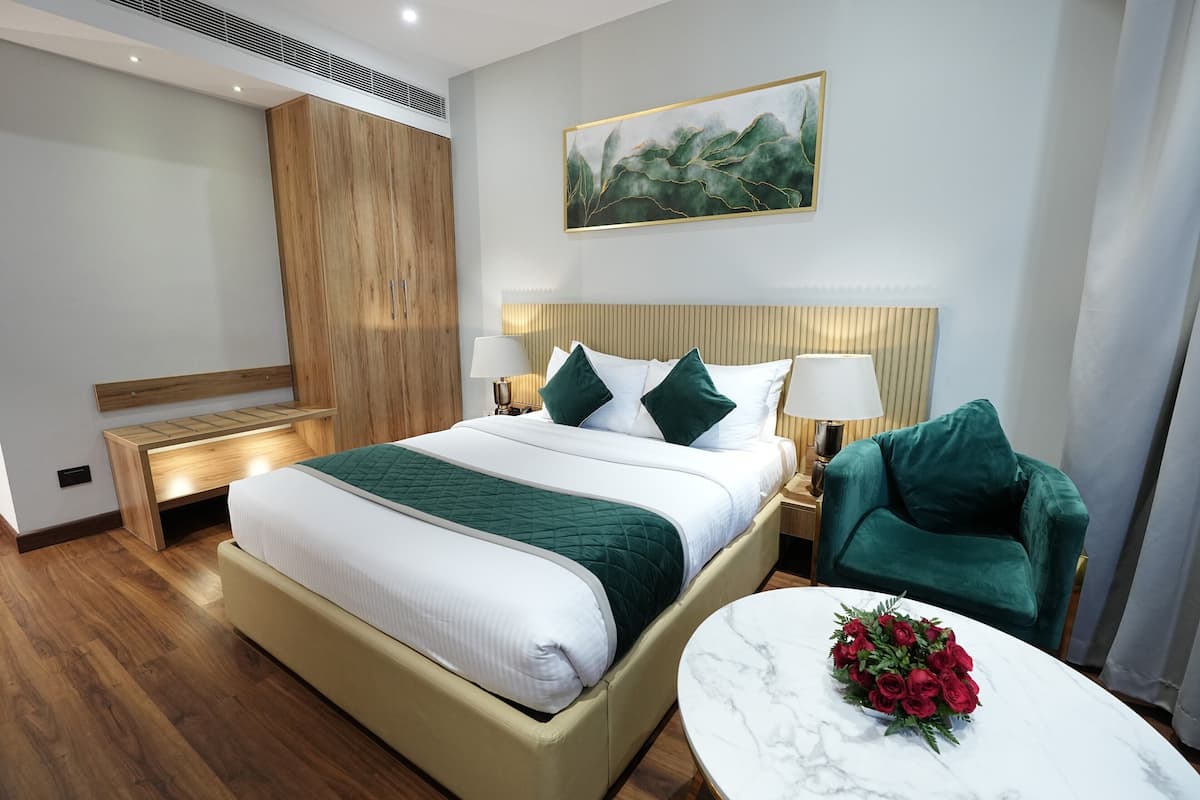 A modern hotel room with a large bed, a wooden wardrobe, a green armchair, a marble table with a bouquet of red roses, and a painting on the wall.
