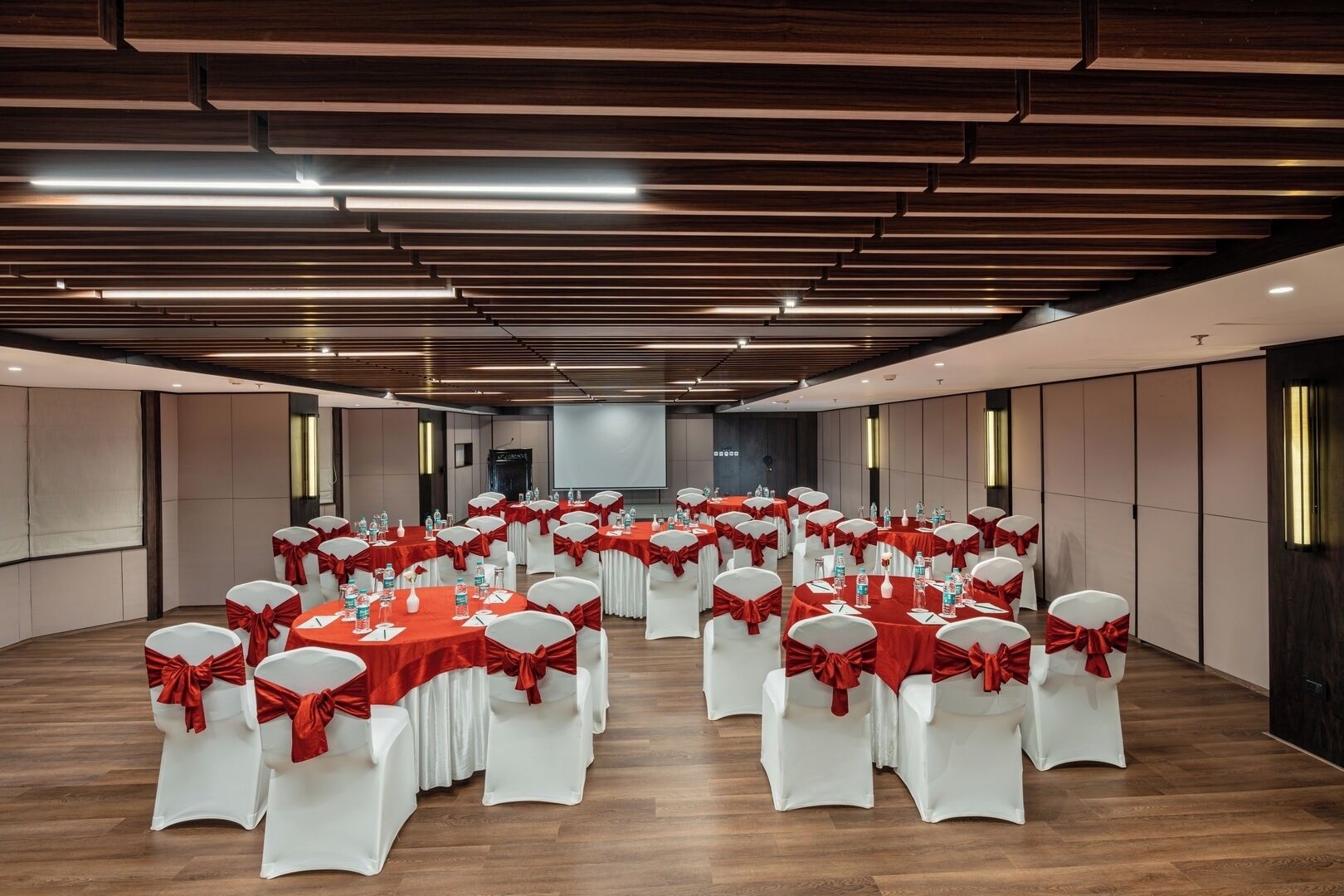 A conference room with rows of tables and chairs, each with red sashes, and a large screen at the front.
