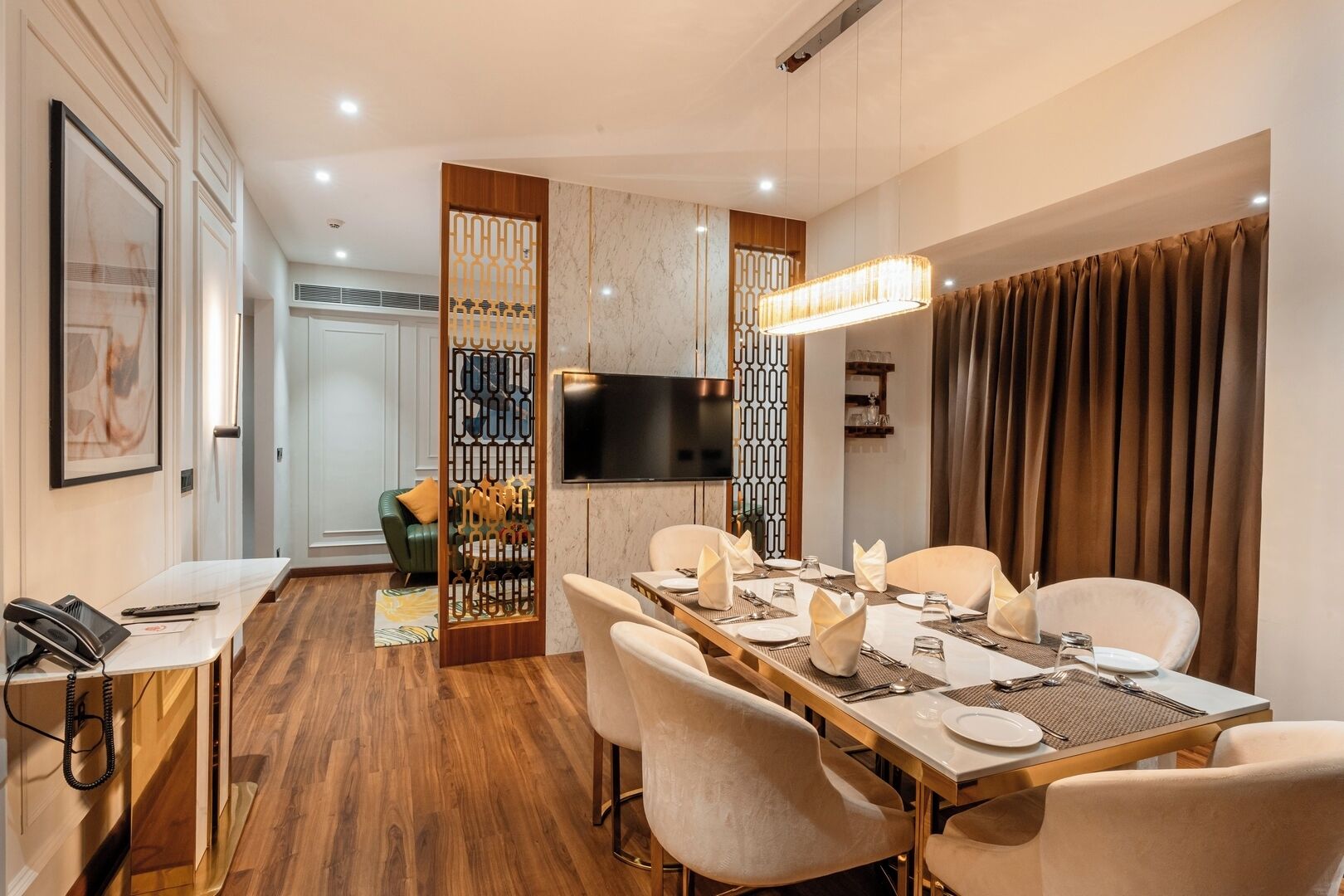 Un comedor con una larga mesa dispuesta para seis personas, rodeada de sillas, un televisor montado en la pared y un separador decorativo con un diseño geométrico.