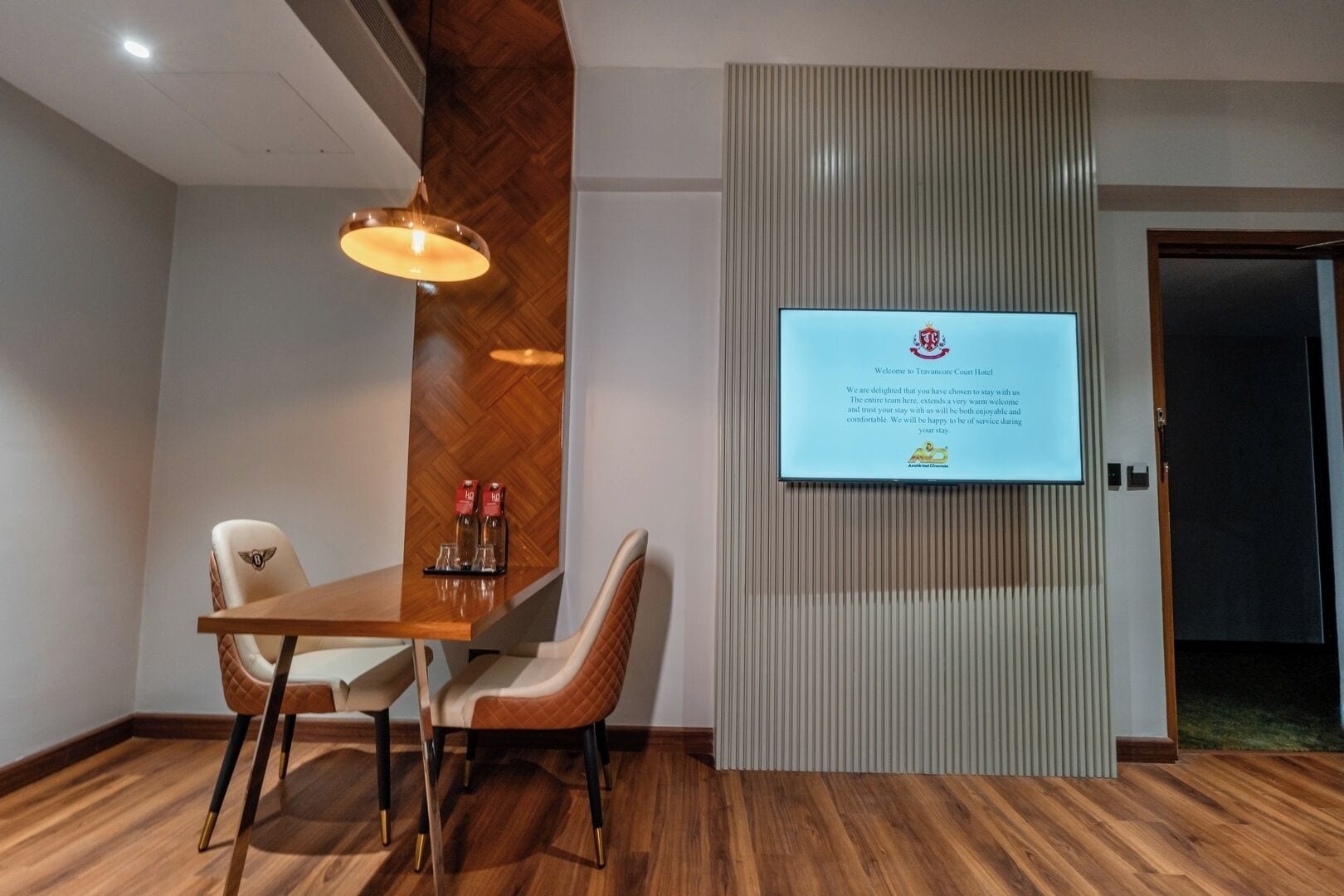 Un'area reception di un hotel con una scrivania in legno, due poltrone, una televisione a parete che mostra un messaggio di benvenuto e un pavimento in legno.