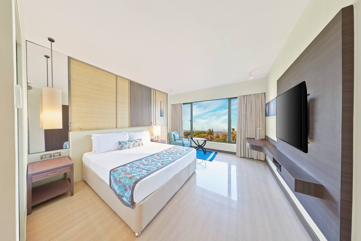A modern hotel room with a large bed, a flat-screen TV, a desk, and a view of the outdoors.