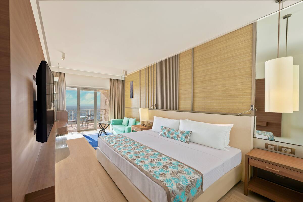 A modern hotel room with a large bed, a desk, a chair, a TV, and a balcony with a view of the ocean.