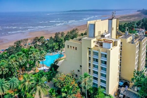 Un hotel con piscina rodeado de palmeras, vista a la playa y un edificio alto con balcones.