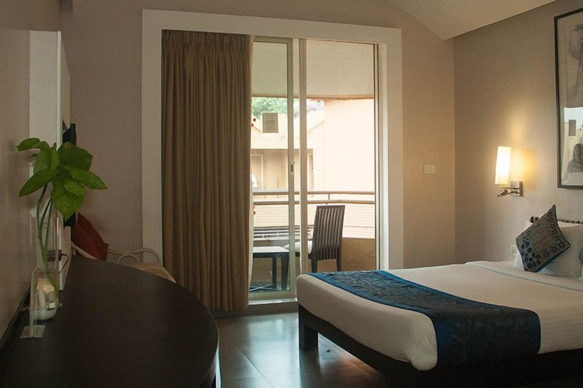 A hotel room with a bed, a desk, a chair, a plant, and a balcony with a table and chairs.