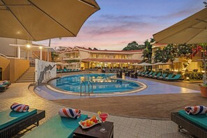 A hotel pool area with lounge chairs, a swimming pool, and a restaurant with outdoor seating. - Whispering Palms Beach Resort (Candolim)
