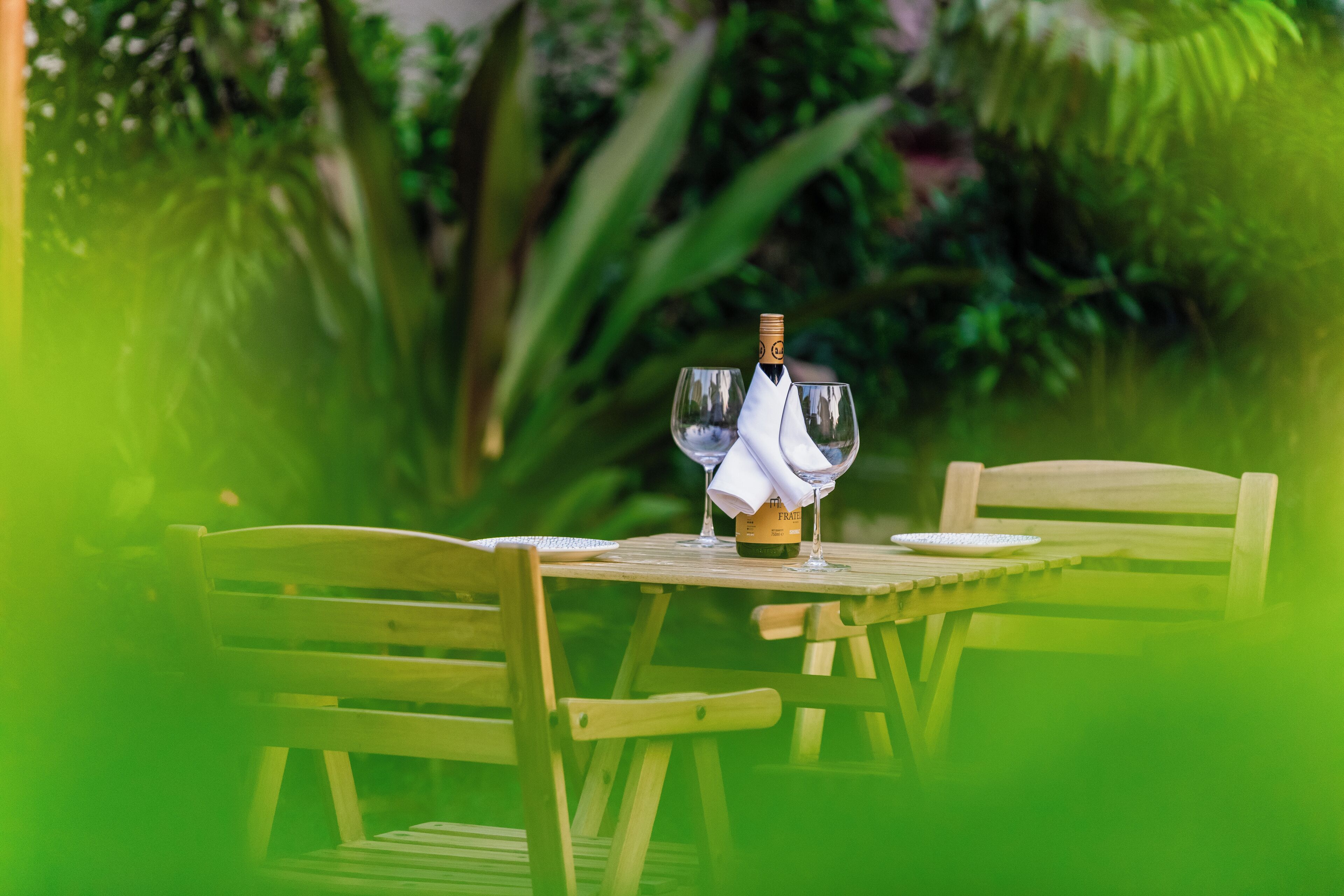 A wooden table with two wine glasses and a bottle of wine, set outdoors with lush greenery in the background.