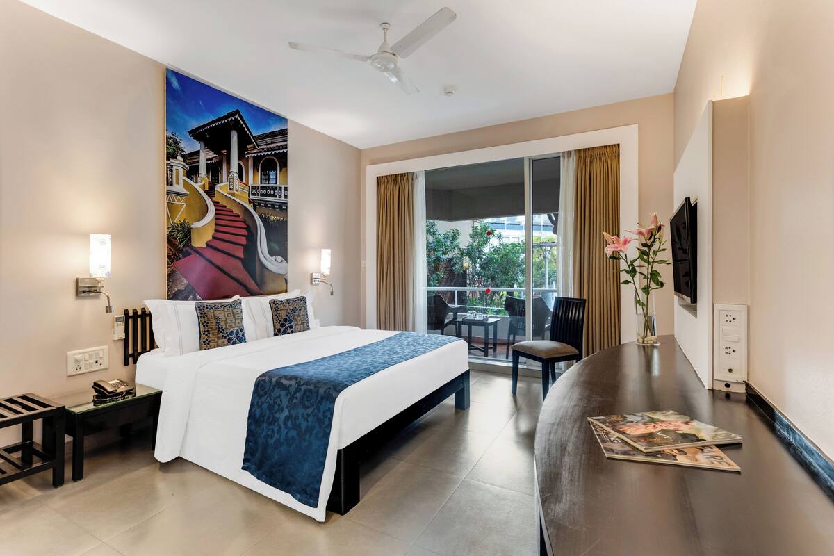 A hotel room with a large bed, a desk, a chair, a balcony with a view, and a ceiling fan.