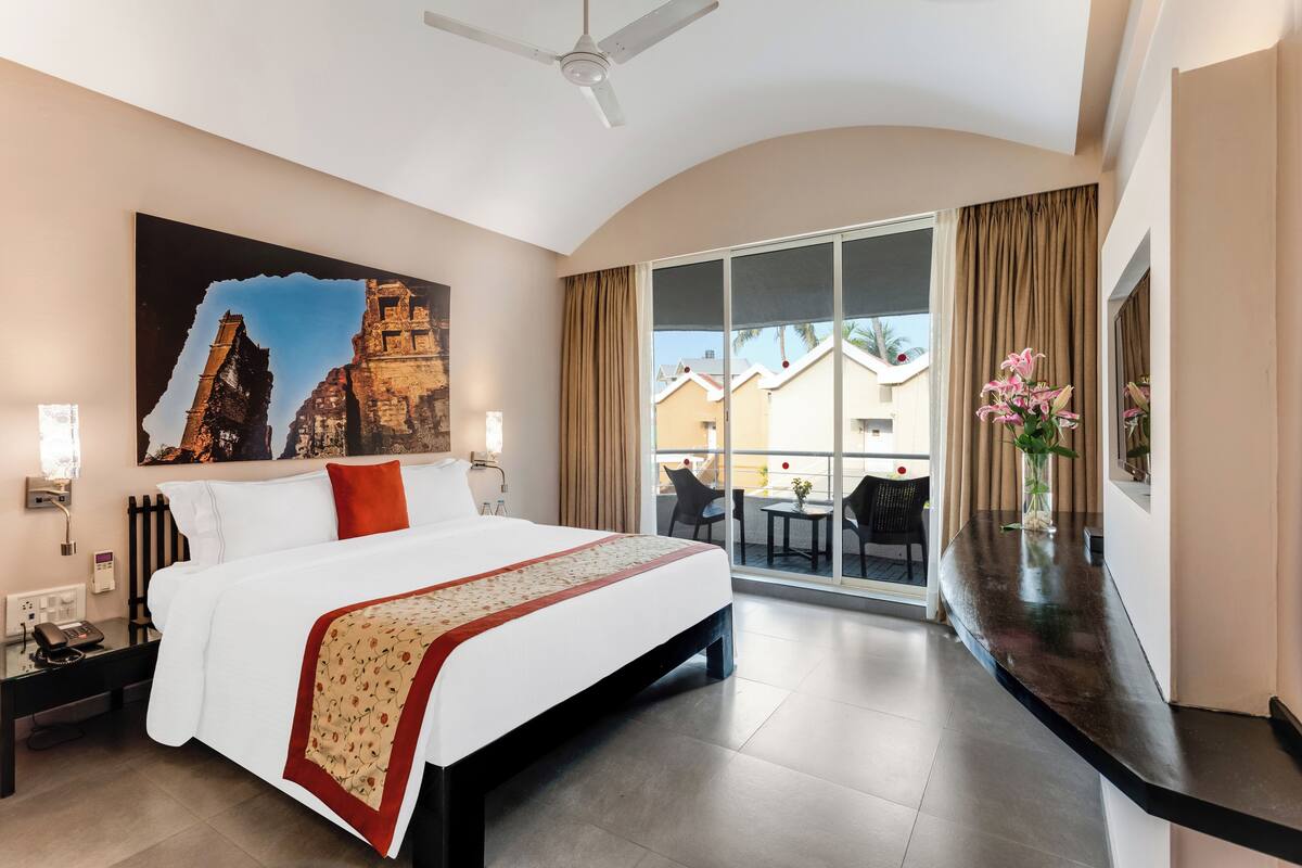 A hotel room with a large bed, a view of the outside, a painting on the wall, and a vase with flowers on the nightstand.