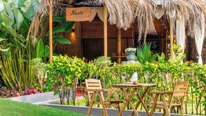 An outdoor seating area with wooden chairs and a table, surrounded by lush greenery and a thatched roof structure. - Whispering Palms Beach Resort (Candolim)