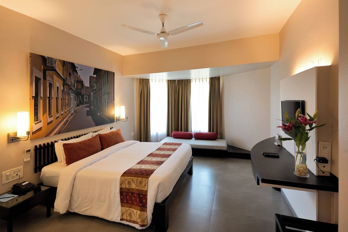 A hotel room with a large bed, a desk, a chair, a ceiling fan, a painting of a street scene, a vase with flowers, and a TV on a stand.