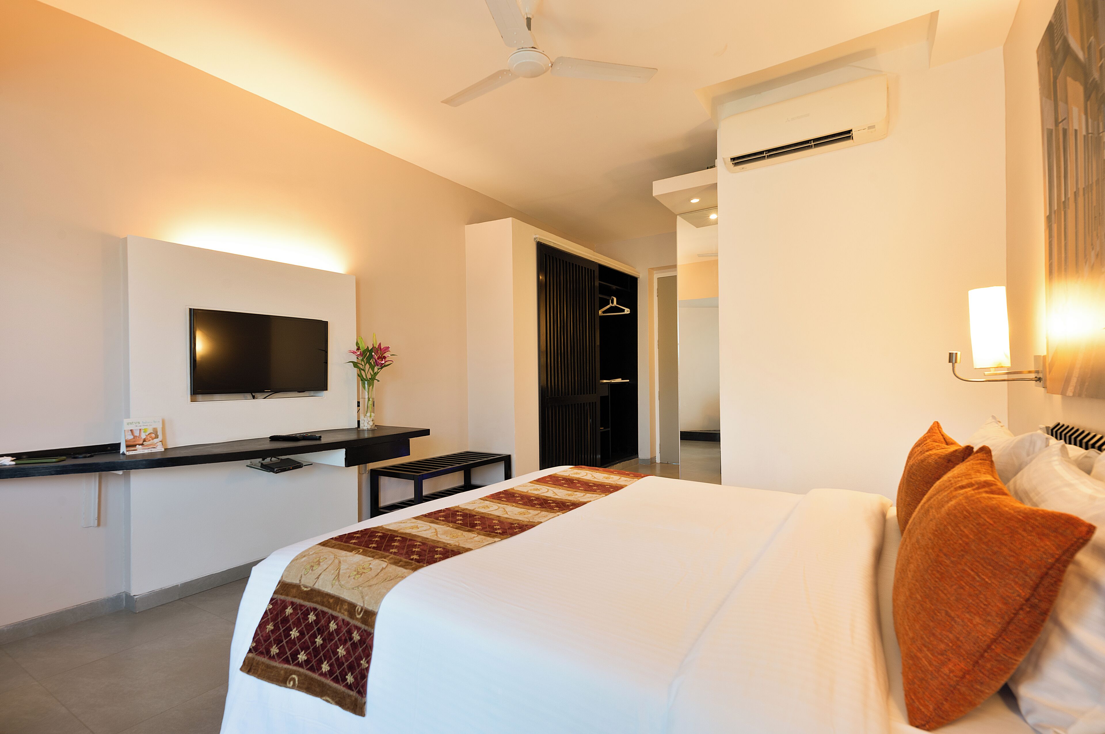A hotel room with a bed, a TV mounted on the wall, a desk with a chair, and a hanging coat rack.