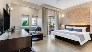 A hotel room with a bed, a desk, a TV, a sofa, and a balcony with plants. - Whispering Palms Beach Resort (Candolim)