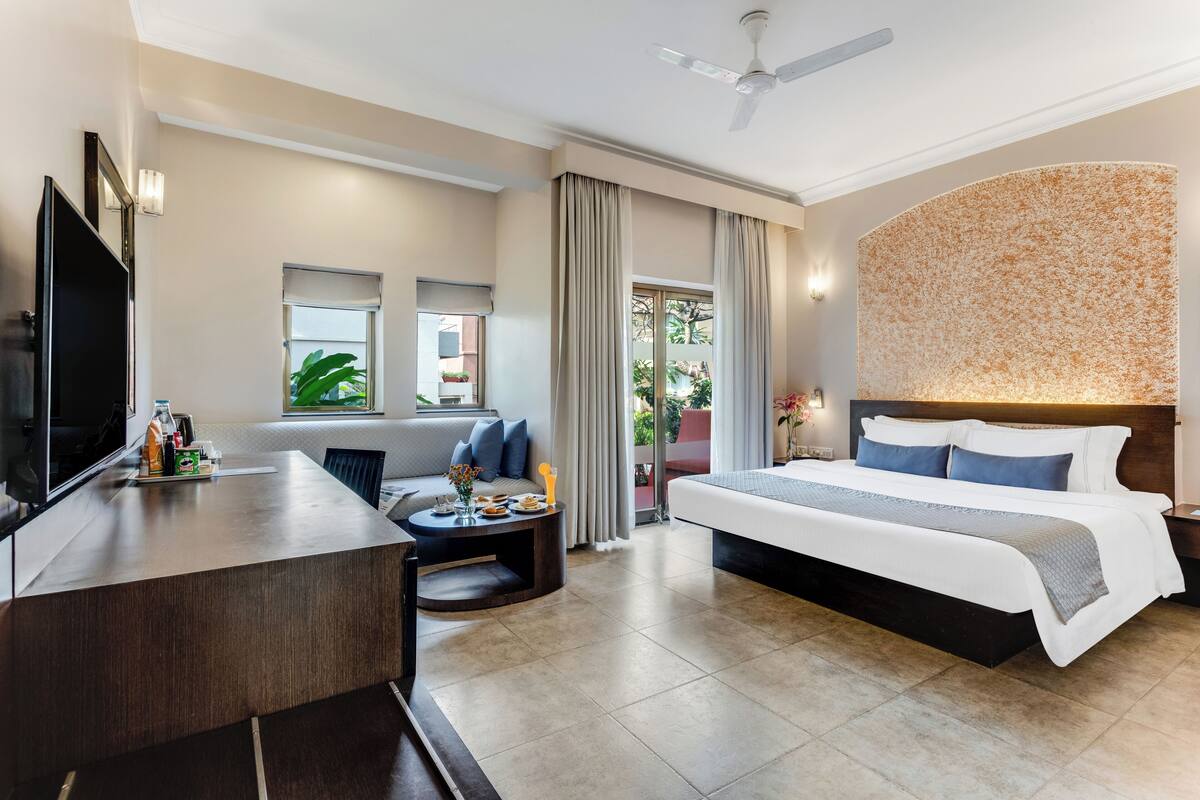 A hotel room with a bed, a desk, a TV, a sofa, and a balcony with plants.