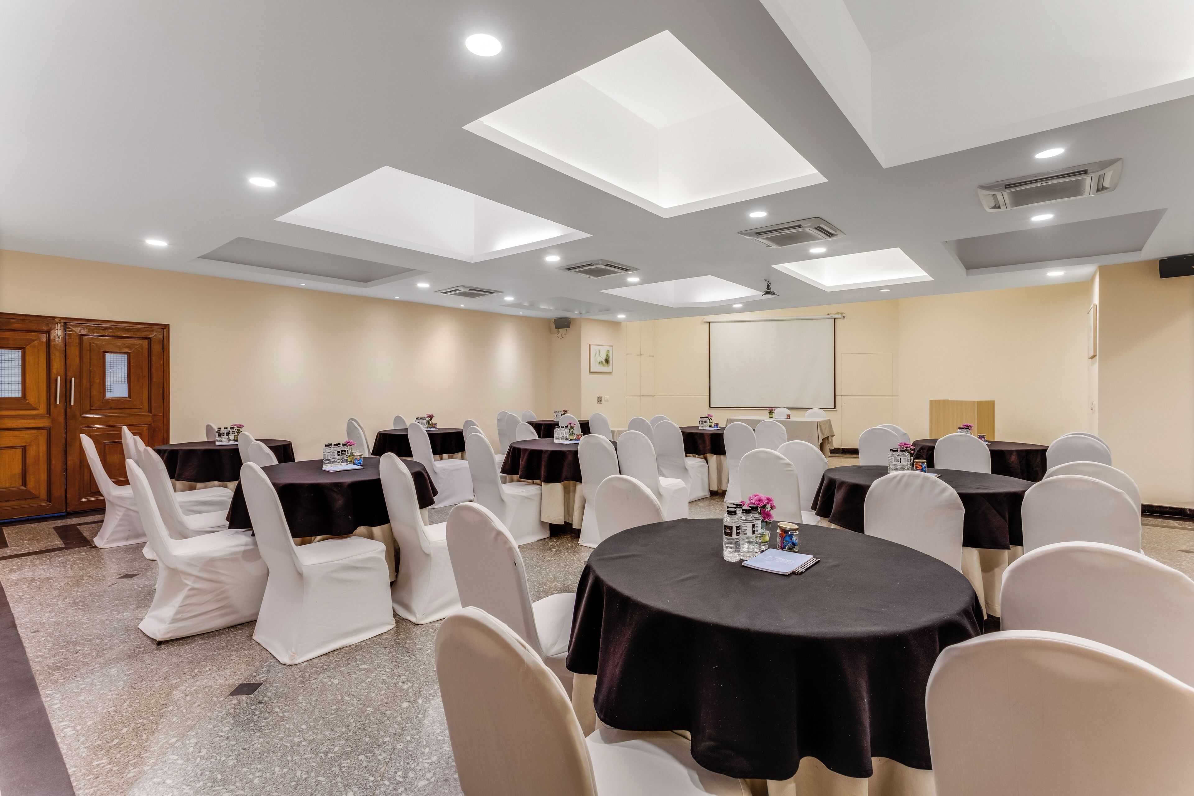 Un salón de conferencias con varias mesas y sillas, equipado con luces en el techo y una pantalla de proyector.