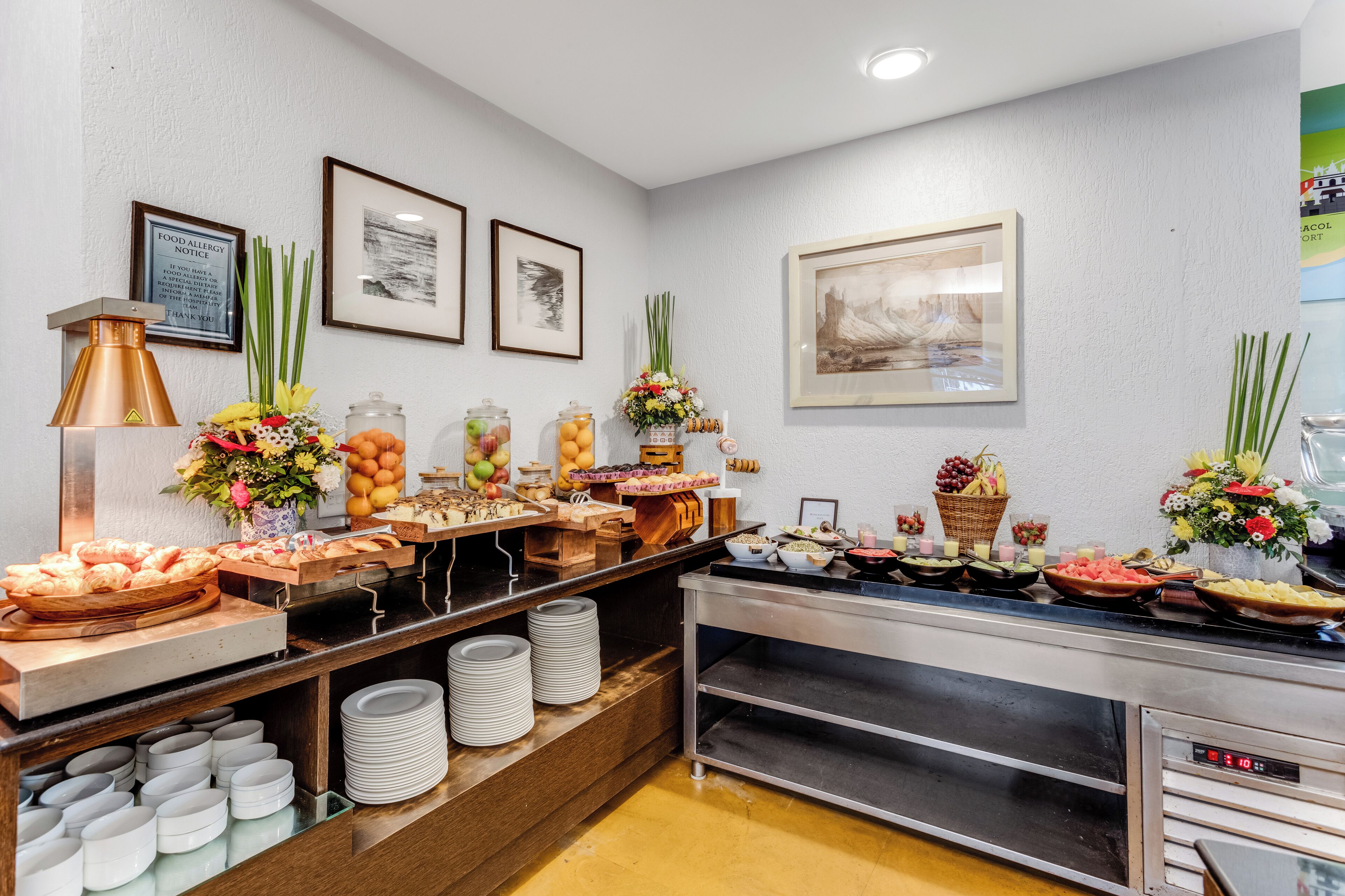 Zona de bufé con diversas opciones de comida, un aviso sobre alergias alimentarias y cuadros en la pared.