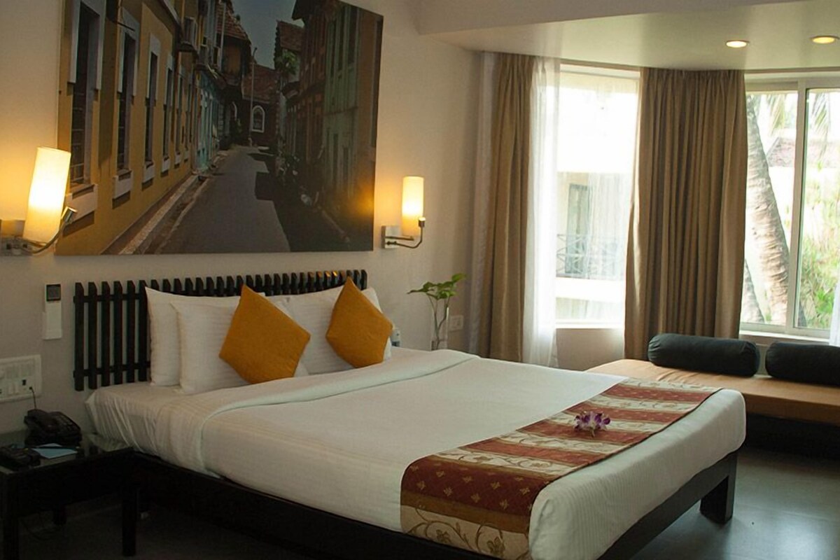 A hotel room with a large bed, two wall-mounted lamps, a window with curtains, a bench with cushions, and a painting of a street scene.