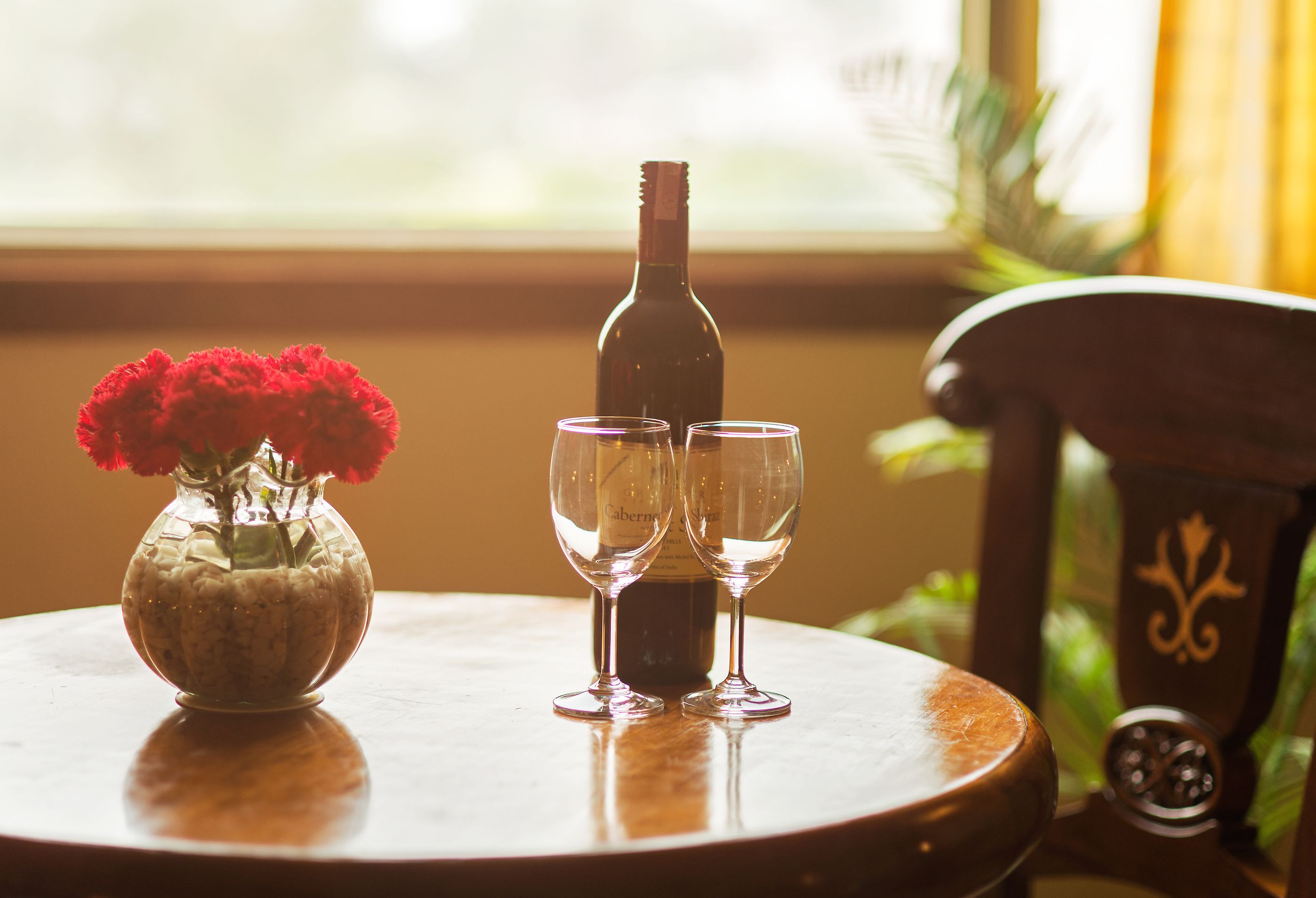 Een tafel met een fles wijn en twee glazen, een vaas met rode bloemen en een stoel met een decoratieve rugleuning.