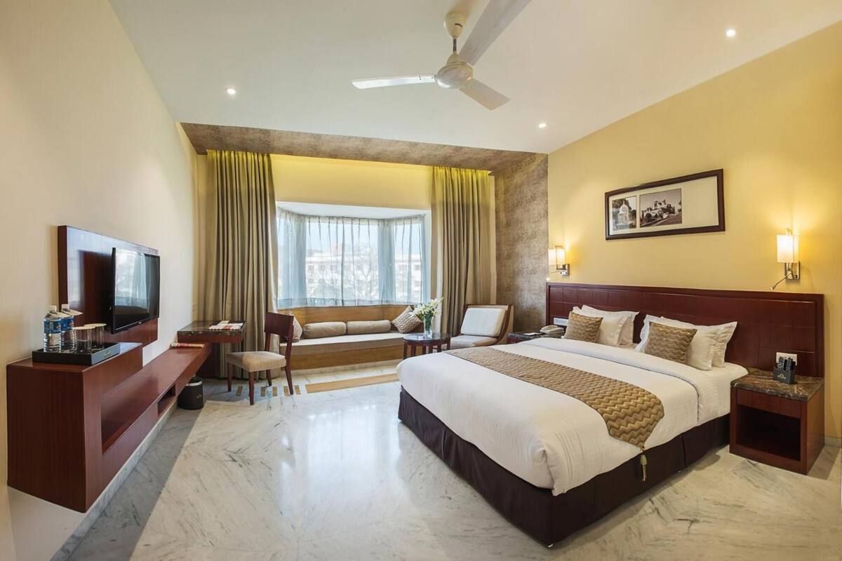 A hotel room with a large bed, a desk with a flat-screen TV, a sofa, and a window with curtains.