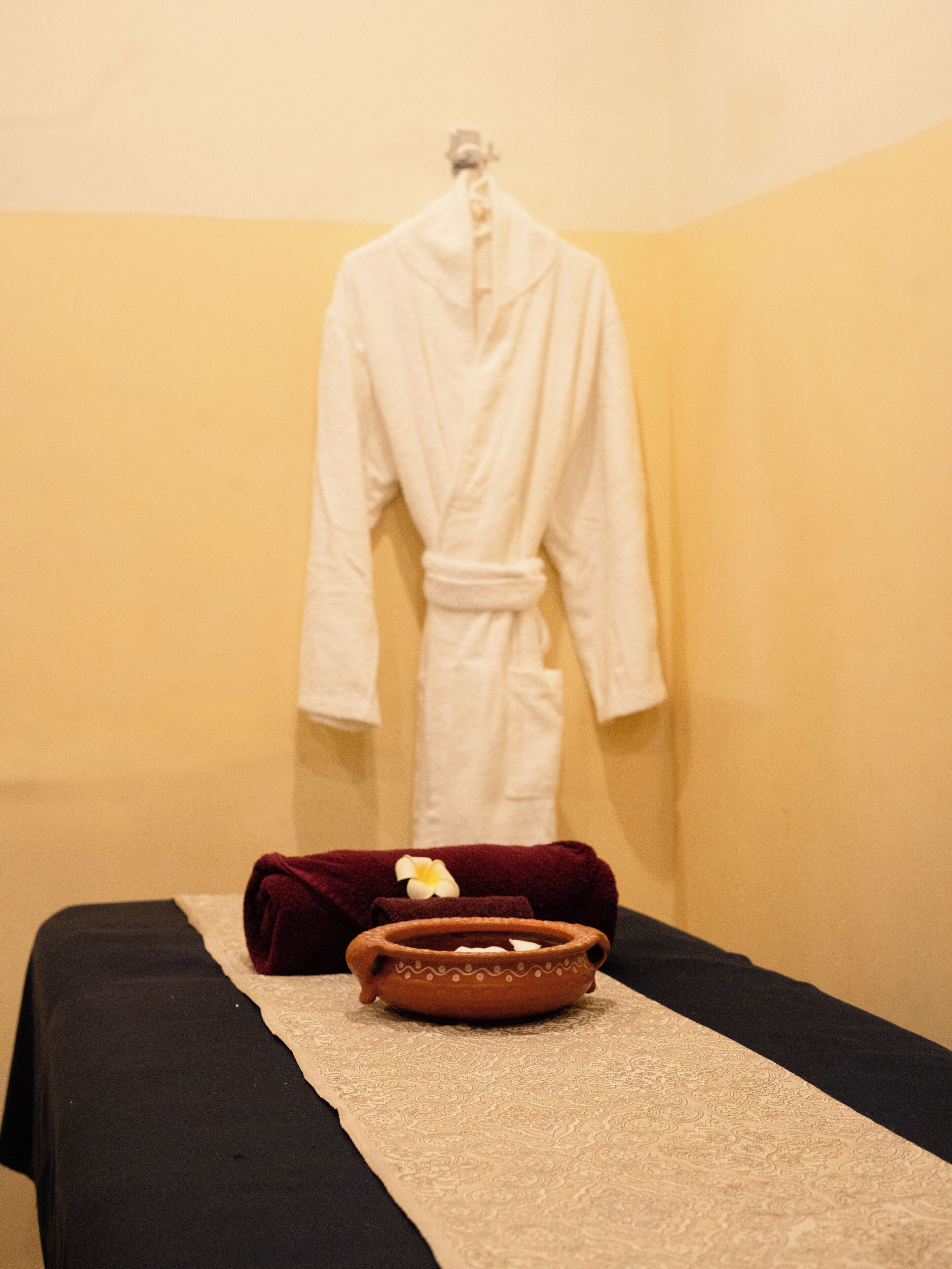 A spa room with a black massage table, a white robe, and a terracotta bowl with a flower.