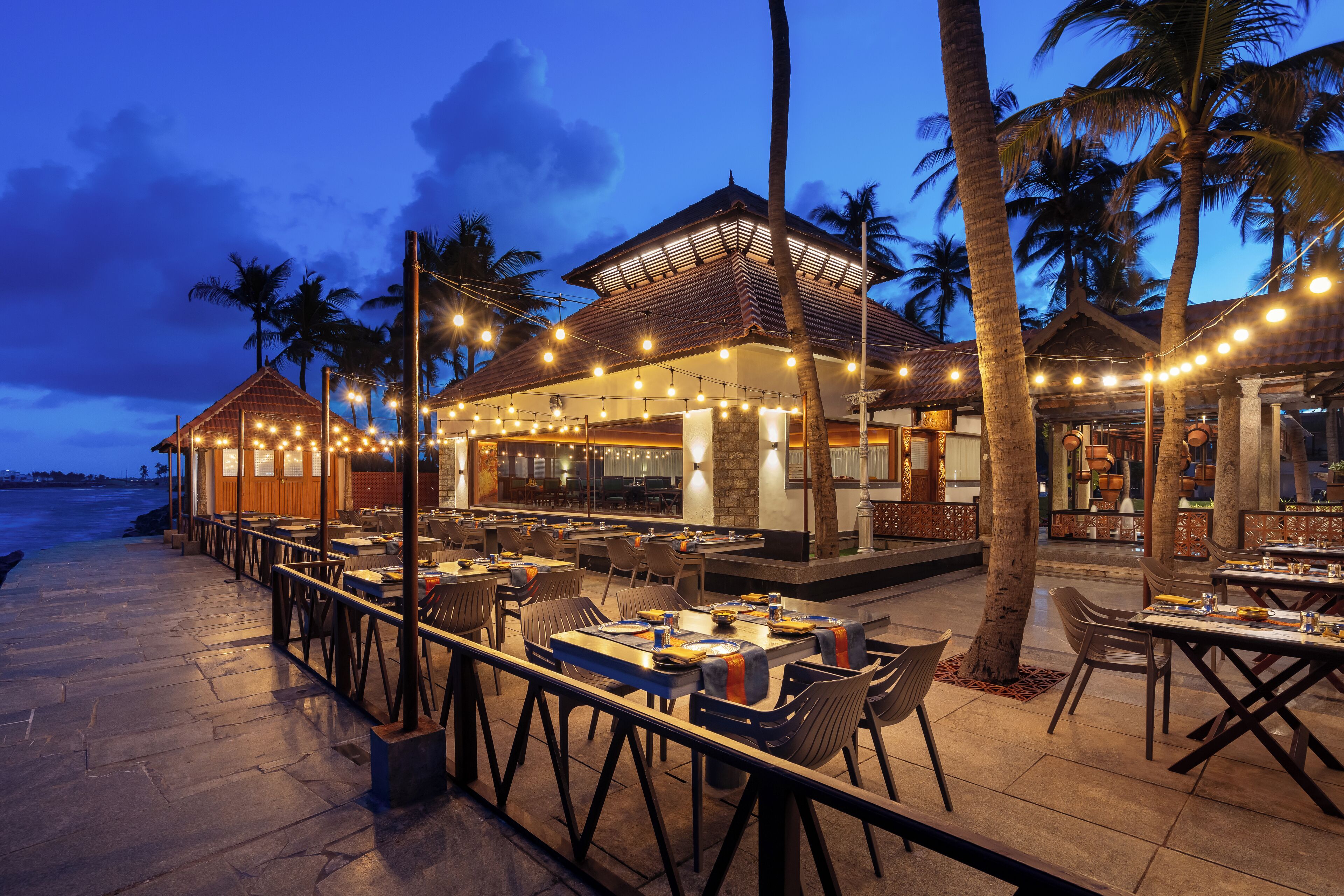 Un restaurante al aire libre con mesas y sillas, palmeras y luces colgantes.