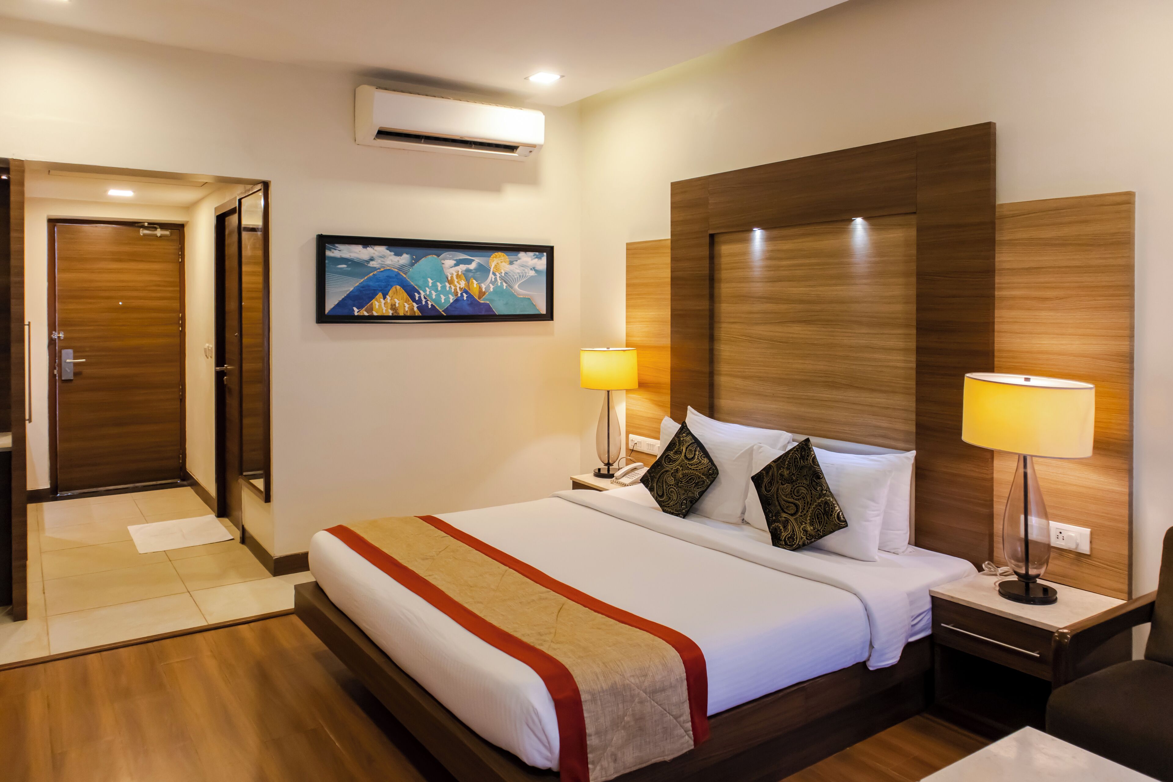A hotel room with a large bed, two bedside lamps, a wooden headboard, a painting on the wall, and an air conditioning unit.