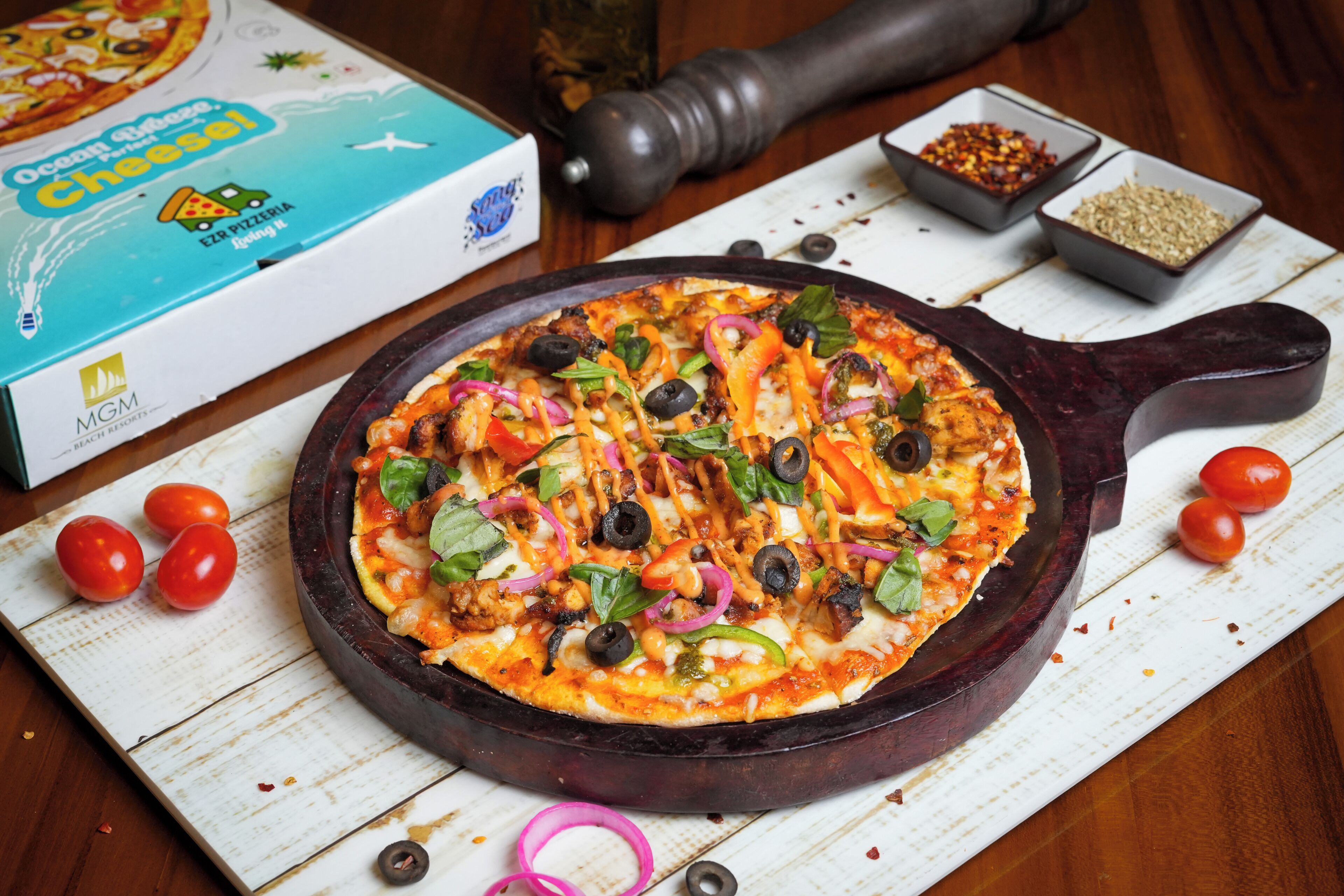 A pizza on a cast iron skillet, surrounded by cherry tomatoes, olives, and a pepper mill on a wooden table.