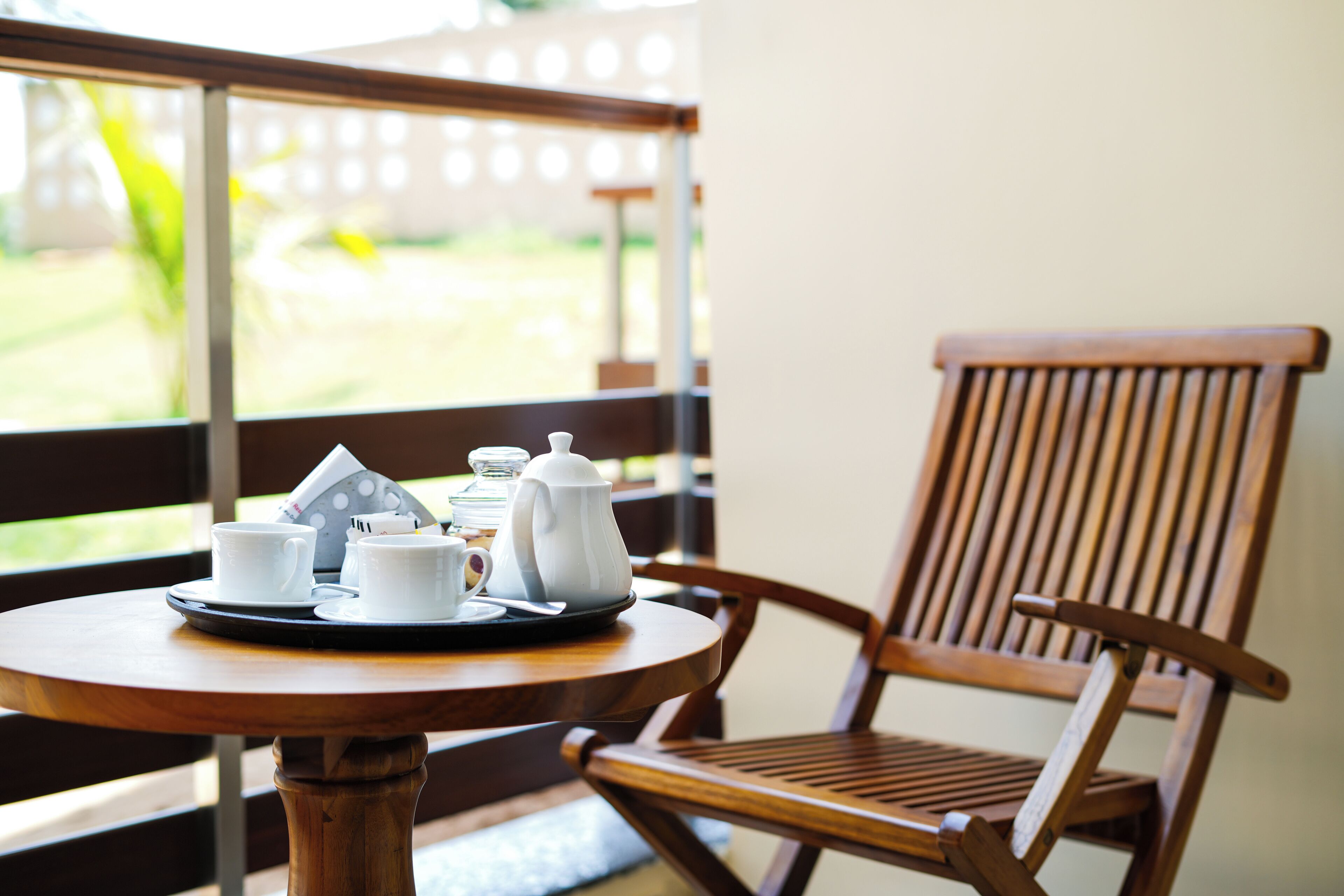 A wooden table with a tray of white cups and a teapot, positioned near a window with a view of greenery.