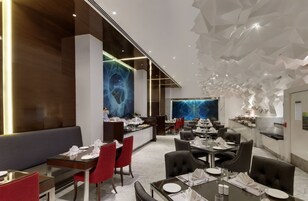 A modern restaurant with a high ceiling and geometric wall design. Tables are set with white tablecloths and red chairs.