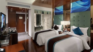 Habitación de hotel con dos camas, un escritorio con computadora, televisor y baño con cabina de vidrio.