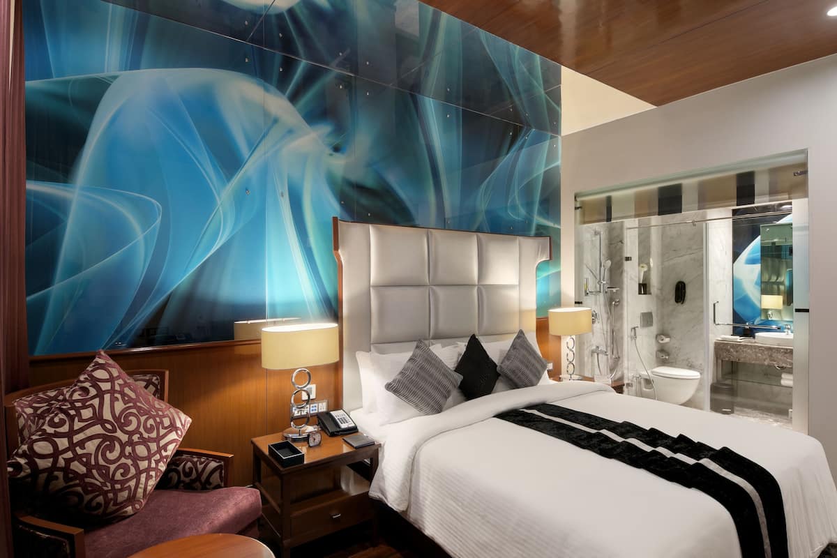 A modern hotel room with a large abstract mural, a plush bed, a sitting area with a chair, and a glass-enclosed shower area.
