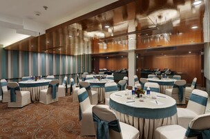 A conference room with multiple round tables, white chairs with teal sashes, and a kitchen area in the background.