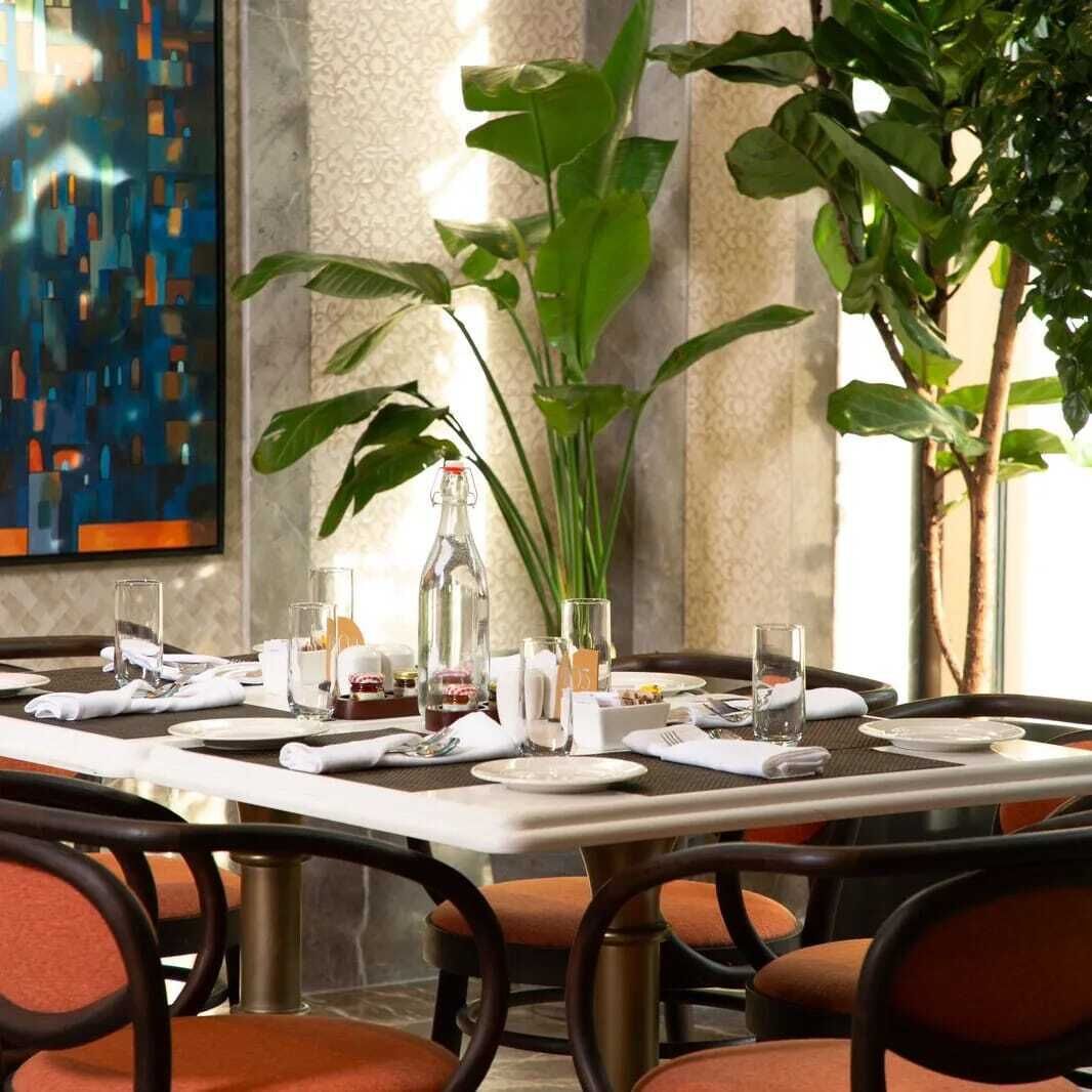 Mesa para cuatro servida con una pintura de la ciudad, plantas verdes grandes y una botella de vino tinto.