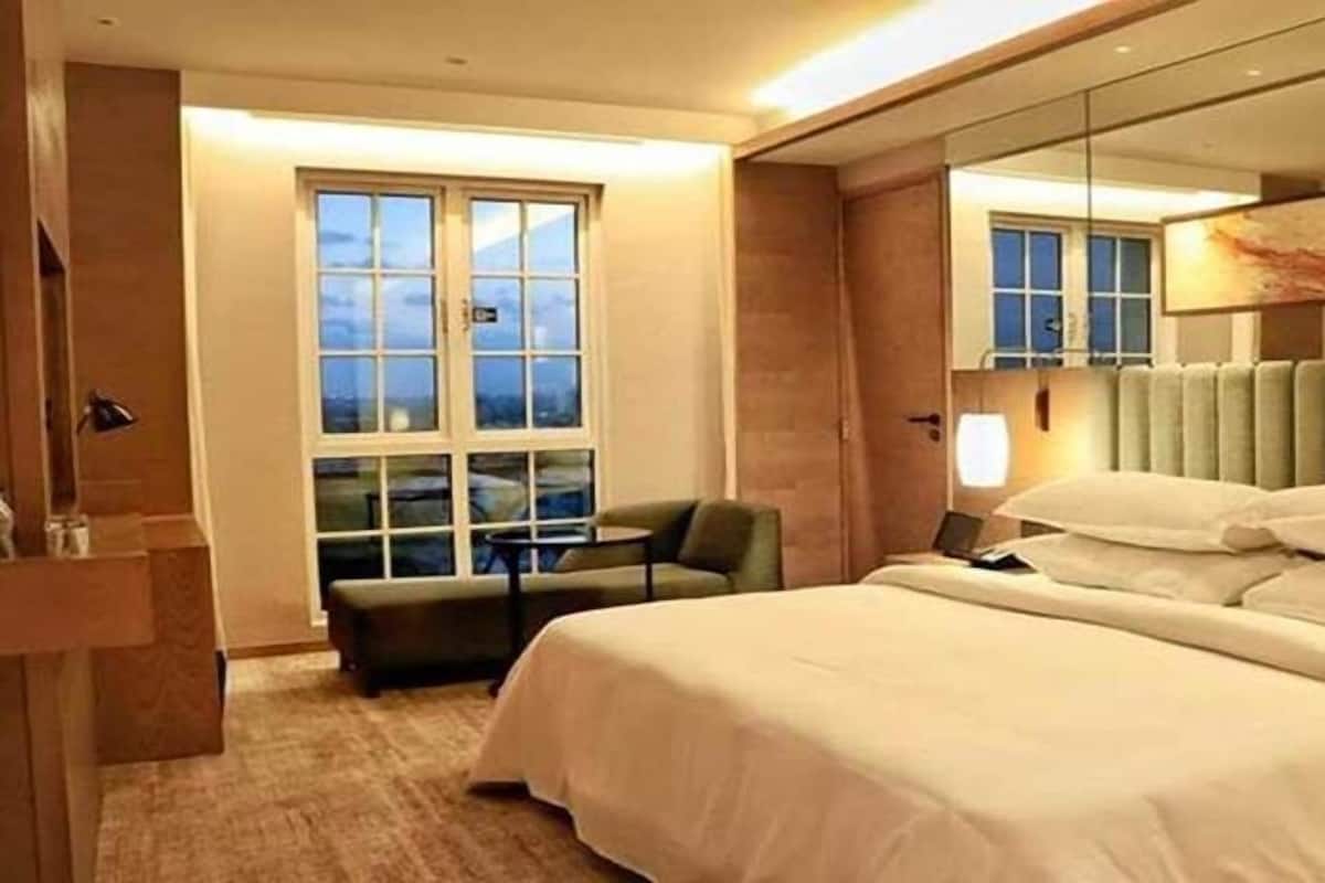 A hotel room with a large bed, a chair, a desk, a window, and a mirror.