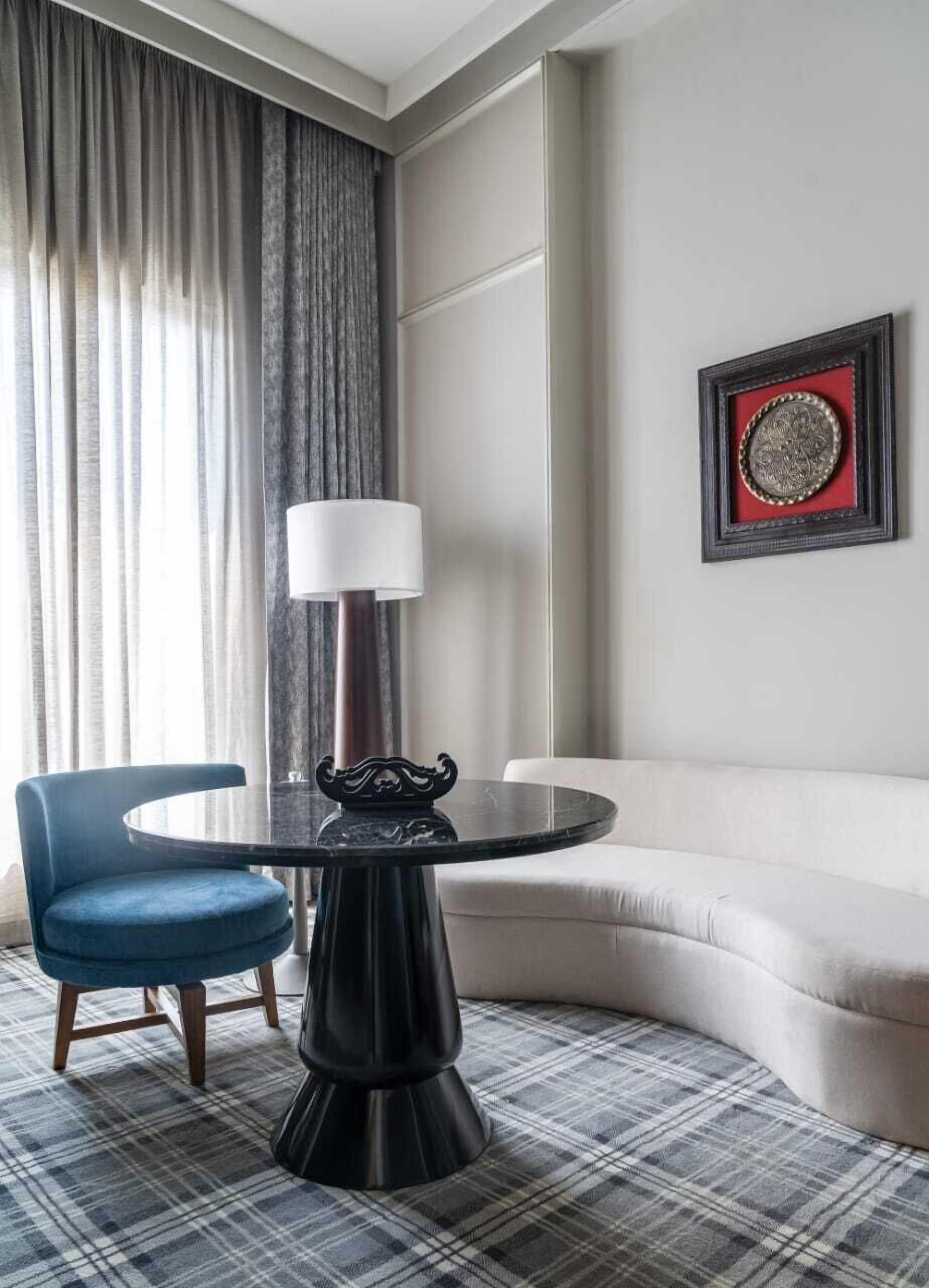 A hotel room with a round black table, a blue chair, a white sofa, a large window with curtains, and a framed picture on the wall.