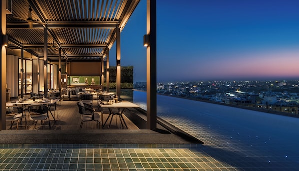 An infinity pool with a city view, outdoor seating area with tables and chairs, and a bar area with a bartender.