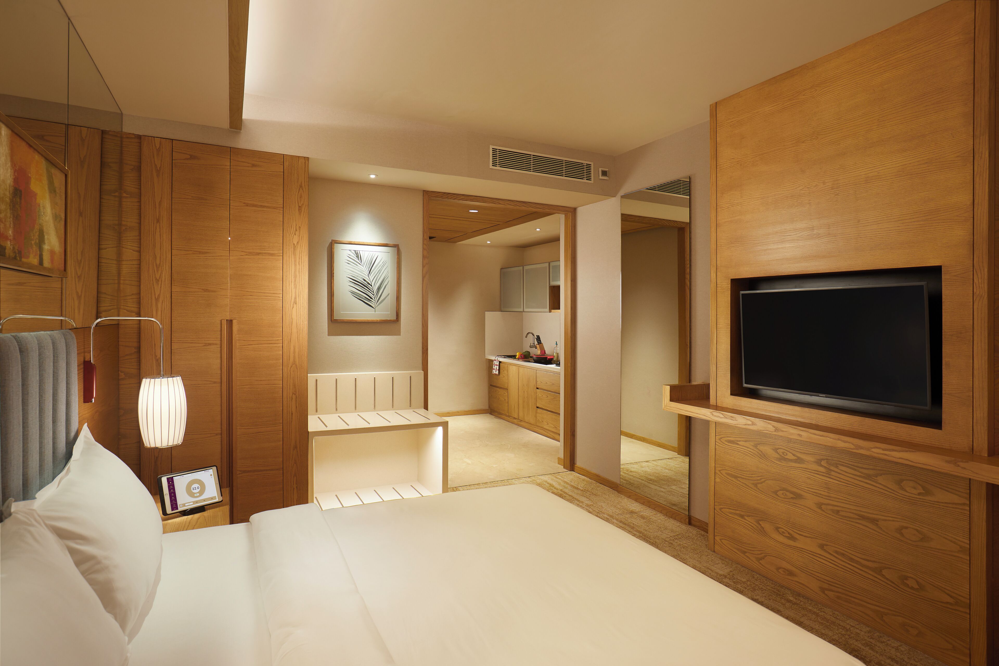 Una habitación de hotel moderna con una pared de acento de madera, un televisor de pantalla plana, una mesita de noche con lámpara y vistas al área de la cocina.