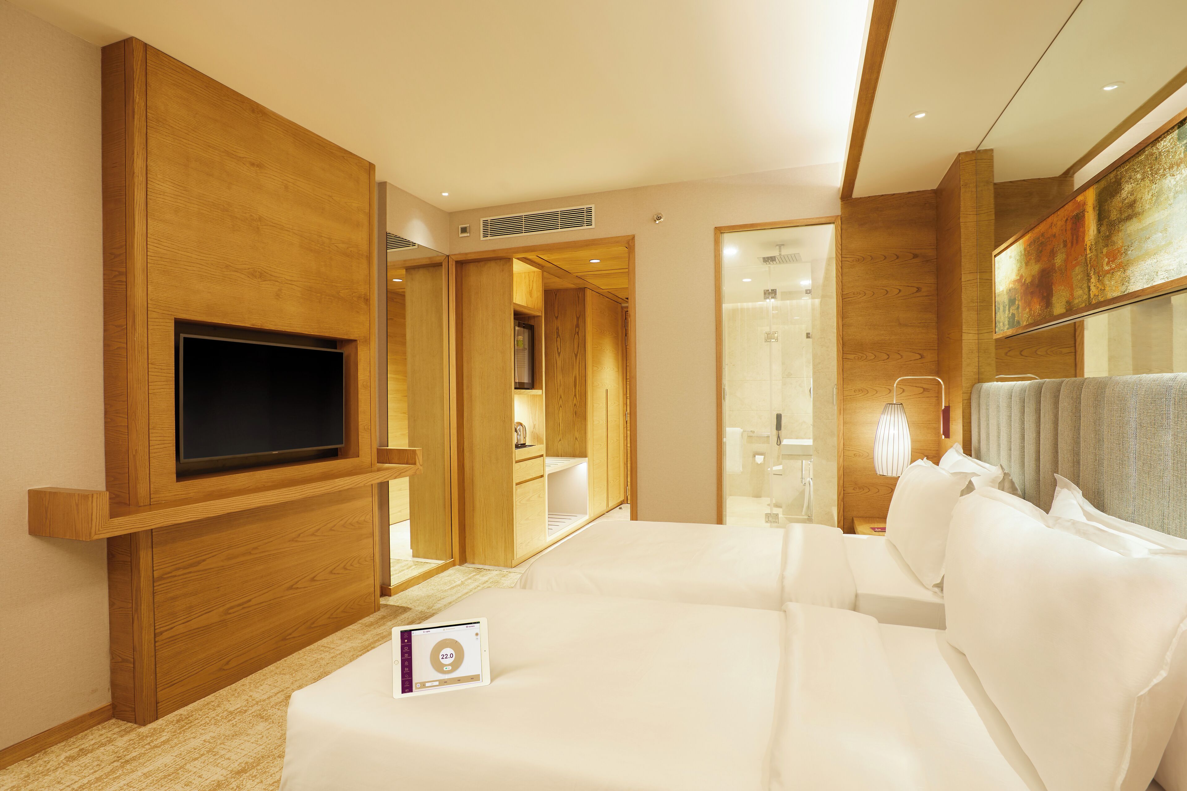 A modern hotel room with a large bed, a flat-screen TV mounted on a wooden panel, a walk-in closet, and a bathroom visible through an open door.