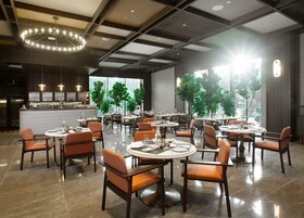 Un restaurante espacioso con mesas redondas y sillas naranjas, ventanas amplias y un diseño moderno en el techo.