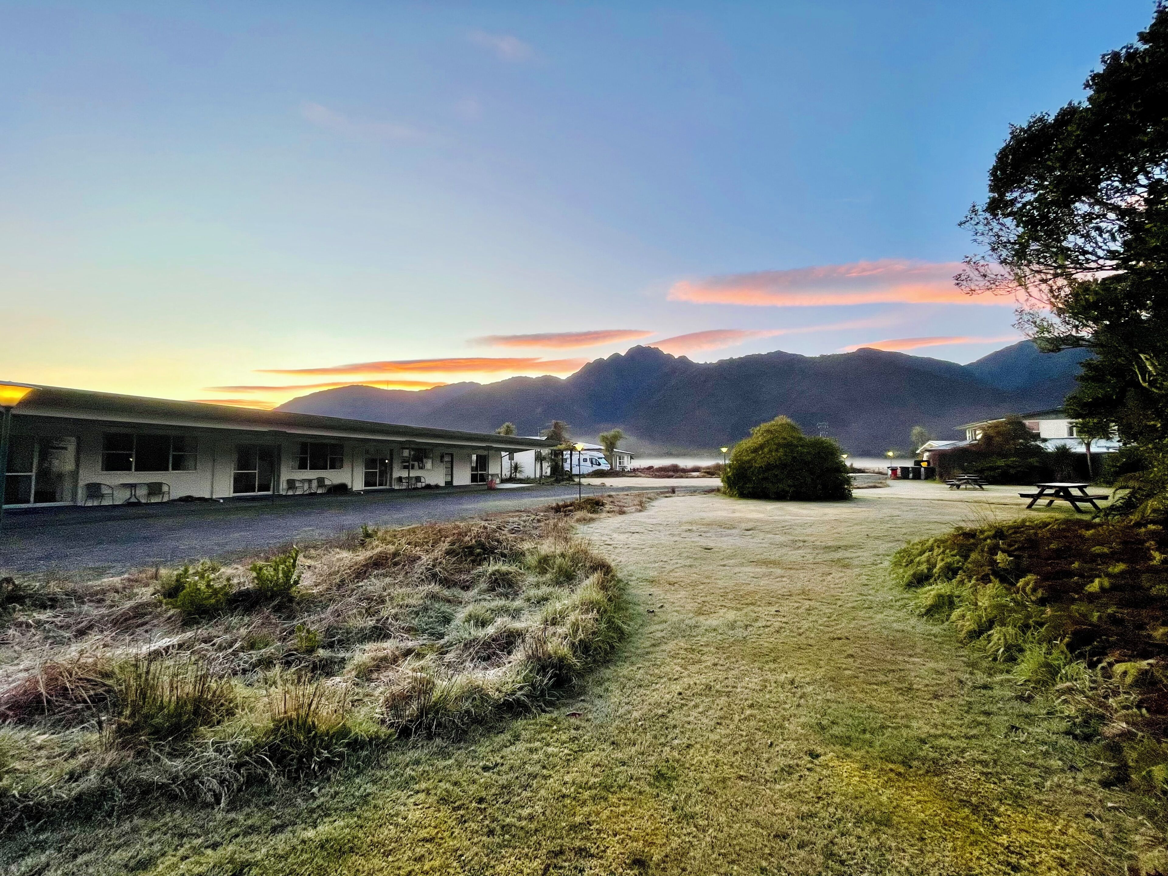 A motel with a simple, single-story structure, a covered parking area, and a view of mountains in the distance.