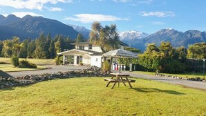 Uma casa com jardim e mesa de piquenique à frente, cercada por montanhas e árvores.