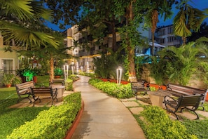 Un camí exterior ben il·luminat amb bancs, plantes en testos i un edifici al fons.